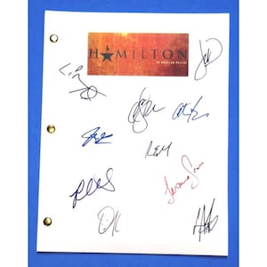 May include: A white sheet of paper with a red, orange, and yellow logo that says "Hamilton: An American Musical". The paper has several blue and black signatures, including "Lin-Manuel Miranda", "Daveed Diggs", and "Anthony Ramos".