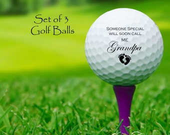 May include: A set of three white golf balls with black text and a footprint design. The text reads "Someone special will soon call me Grandpa".