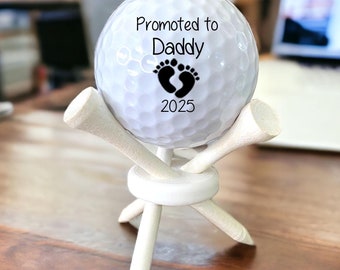 May include: White golf ball with black text and footprint design that reads "Promoted to Daddy 2025" on a white golf ball tee stand.