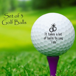 May include: Set of three white golf balls with a black inscription that reads "It takes a lot of balls to say "I do"". The golf balls are on a purple tee and sitting in green grass.