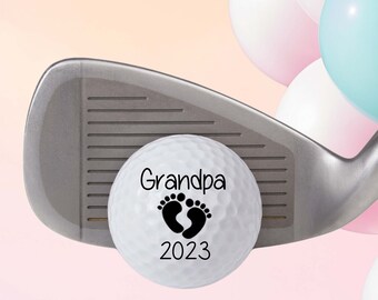 May include: A white golf ball with black text that reads "Grandpa" and a footprint symbol, and the year "2023". The golf ball is resting on a silver golf club with a pink and blue balloon background.