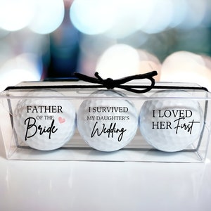 May include: Three white golf balls in a clear plastic box with a black ribbon. The golf balls are decorated with text. The first golf ball says "Father of the Bride" with a pink heart. The second golf ball says "I survived my daughter's wedding". The third golf ball says "I loved her first".