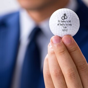 May include: White golf ball with black text that reads "It takes a lot of balls to say "I do" 10.29.22" and a drawing of two wedding rings.