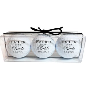 May include: Three white golf balls in a clear plastic box with a black ribbon. Each ball has the text "Father of the Bride" and the date "9.9.2024" printed on it.