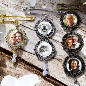 May include: Three gold and silver tone photo charm brooches with pearl accents. Each brooch features a different photo of a person, pet, or couple. The brooches are displayed on a wooden surface with white flowers.