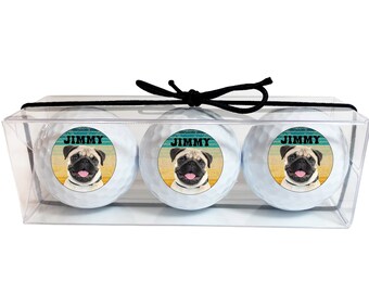 May include: Three white golf balls with a retro-style image of a pug dog and the name "JIMMY" printed on each ball. The golf balls are in a clear plastic box with a black ribbon.