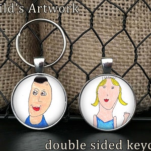 May include: Double-sided keychain featuring two round, silver-colored charms with colorful drawings of children's artwork. The text "Your Child's Artwork" is at the top of the image.