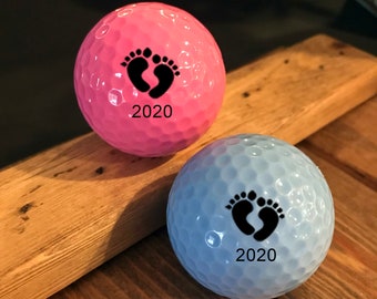 May include: Two golf balls, one pink and one light blue, with black baby footprints and the year 2020 printed on them.