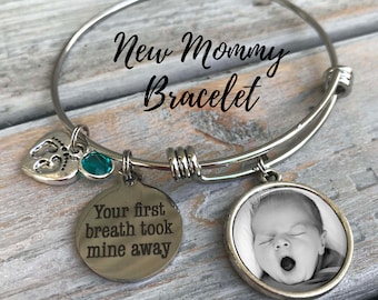 May include: A silver charm bracelet with the text "New Mommy Bracelet" on the bangle. The bracelet has three charms: a heart with a footprint, a turquoise gemstone, and a round charm with a black and white photo of a baby. The round charm has the text "Your first breath took mine away" engraved on the back.