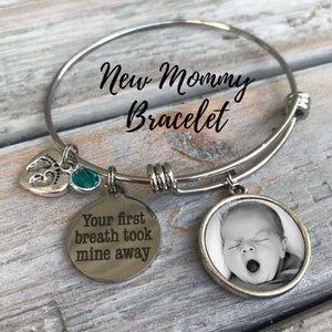 May include: A silver charm bracelet with the text "New Mommy Bracelet" on the bangle. The bracelet has three charms: a heart with a footprint, a turquoise gemstone, and a round charm with a black and white photo of a baby. The round charm has the text "Your first breath took mine away" engraved on the back.