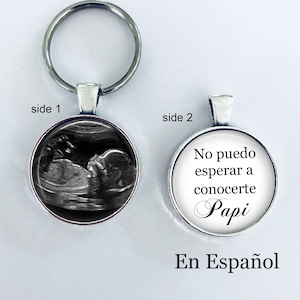 May include: A silver keychain with a round charm on each side. One side features a black and white ultrasound image of a baby. The other side has the text "No puedo esperar a conocerte Papi En Español".