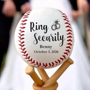 May include: A white baseball with red stitching and the text "Ring Security" with a wedding ring icon and the name "Benny" and the date "October 3, 2026". The baseball is on a wooden stand made of two crossed baseball bats.