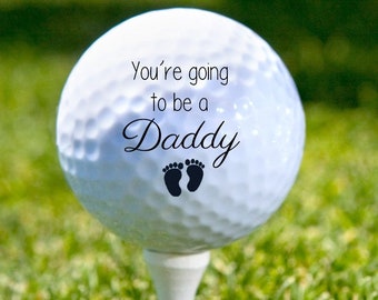 May include: A white golf ball with the text "You're going to be a Daddy" and a pair of black footprints on a green grassy surface.