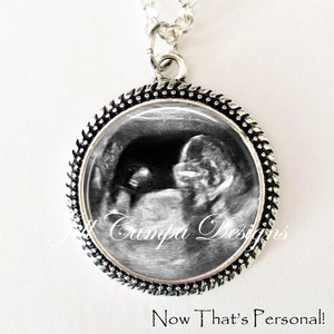 May include: A silver necklace with a round pendant featuring a black and white ultrasound image of a baby. The pendant has a silver beaded border. The text "Now That's Personal!" is printed below the pendant.