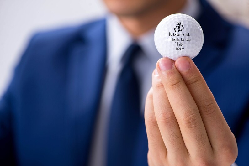 May include: White golf ball with black text that reads "It takes a lot of balls to say "I do" 10.29.22" and a drawing of two wedding rings.