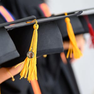 May include: A black graduation cap with a yellow tassel and a small round photo charm attached. The photo charm features a person's face.