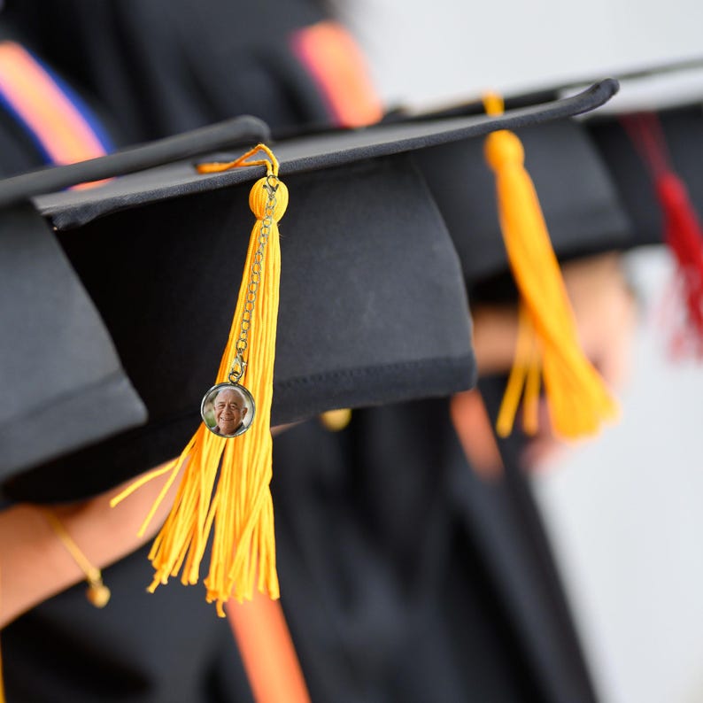 May include: A black graduation cap with a yellow tassel and a small round photo charm attached. The photo charm features a person's face.