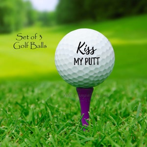 May include: A white golf ball with the text "Kiss My Putt" printed on it. The golf ball is on a purple tee and is sitting on green grass.