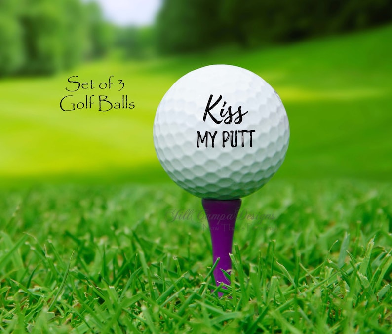 May include: A white golf ball with the text "Kiss My Putt" printed on it. The golf ball is on a purple tee and is sitting on green grass.