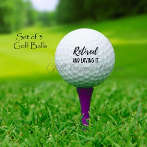 May include: Set of three white golf balls with black text that reads "Retired and Loving It". The golf balls are on a green tee.