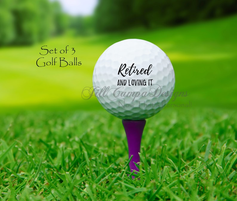 May include: Set of three white golf balls with black text that reads "Retired and Loving It". The golf balls are on a green tee.