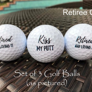 May include: Set of three white golf balls with black lettering. The golf balls say "Retired and Loving It", "Kiss My Putt", and "Retired and Loving It".