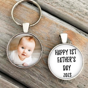 May include: A silver keychain with a round photo charm featuring a baby and a round charm with the text "HAPPY 1ST FATHER'S DAY 2023".