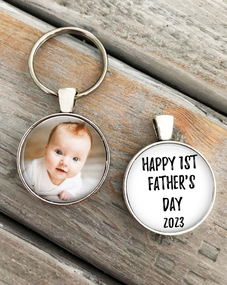 May include: A silver keychain with a round photo charm featuring a baby and a round charm with the text "HAPPY 1ST FATHER'S DAY 2023".