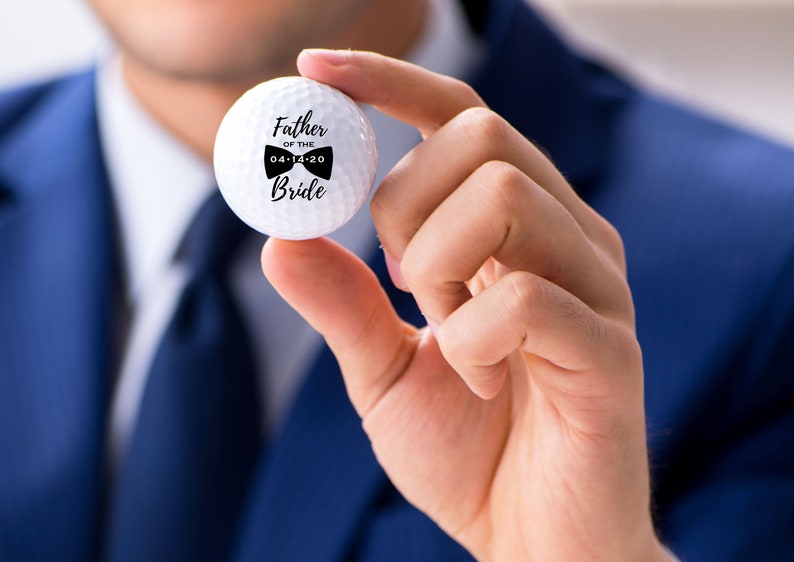 May include: White golf ball with black text that reads "Father of the Bride" and a bow tie graphic with the date "04-14-20".