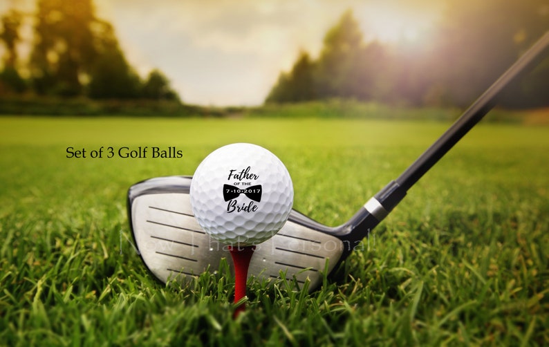May include: Set of three white golf balls with black text and a bow tie design. The text reads "Father of the Bride" with the date "7-10-2017".