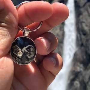 以下が含まれることがあります： 黒と白の超音波画像が写った丸い透明なチャームが付いたシルバーのキーホルダーです。