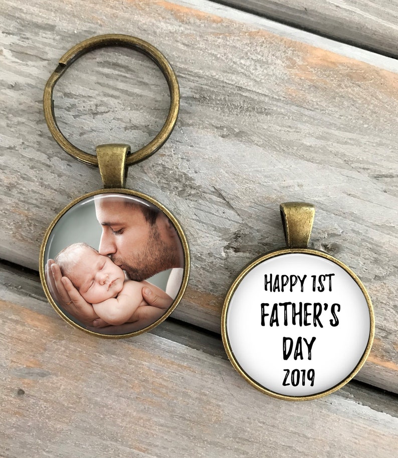 May include: A bronze-colored keychain with a round photo charm featuring a father kissing a baby. The second charm is round and has the text "Happy 1st Father's Day 2019" in black.