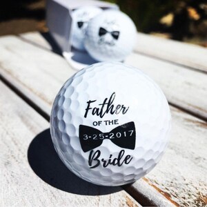 May include: White golf ball with black text and a bow tie design. The text reads "Father of the Bride" and "3-25-2017".