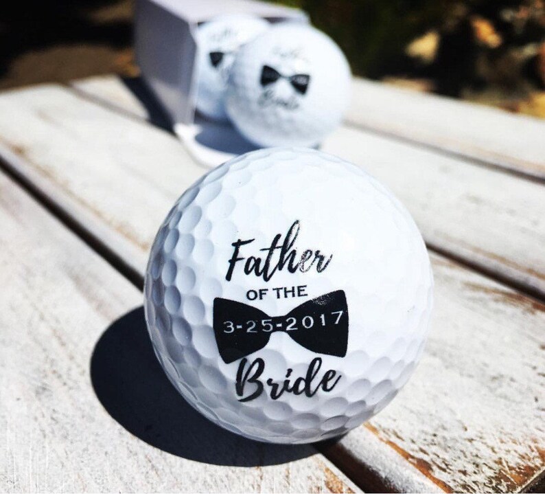 May include: White golf ball with black text and a bow tie design. The text reads "Father of the Bride" and "3-25-2017".