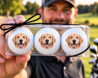 May include: A clear rectangular plastic box containing three white golf balls. Each golf ball features a printed image of a golden retriever dog face with a smiling expression, pink tongue, and dark eyes. The box is tied with a black ribbon.