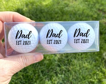 May include: Three white golf balls with black lettering that reads "Dad EST 2021" in a clear plastic box.