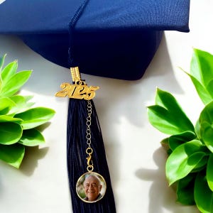 May include: A black graduation tassel with a gold 2025 charm and a round photo charm. The photo charm features a man with white hair and a smile.