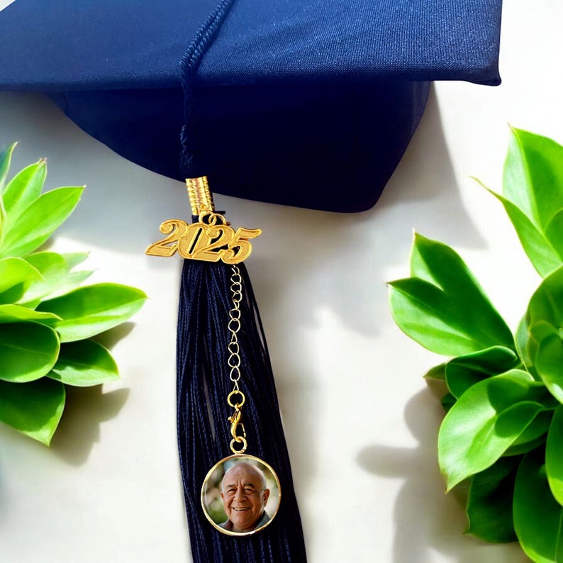 May include: A black graduation tassel with a gold 2025 charm and a round photo charm. The photo charm features a man with white hair and a smile.