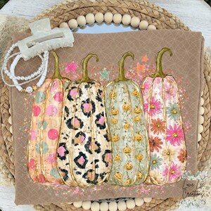 Puede incluir: Una camiseta color canela con un gráfico de cuatro calabazas con patrones distintos: lunares rosas, estampado de leopardo, pequeñas calabazas de Halloween y margaritas coloridas. El diseño incluye estrellas y brillos, junto a una pinza blanca.