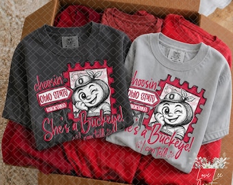 Pode incluir: Uma t-shirt cinzento-escuro e uma cinzento-claro, cada uma com o gráfico de uma personagem de desenho animado com um chapéu com o logótipo O. O design inclui uma borda de selo, o texto Ohio State e a frase choosin' she's a Buckeye.