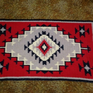 May include: A red, white, and blue woven rug with a geometric pattern. The rug has a square shape and a border of dark blue.