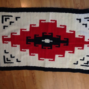 May include: A white crocheted rug with a red, black, and white geometric design. The rug has a navy blue border.