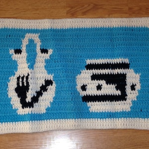 May include: A crocheted rug with a blue background and white border. The rug features two white crocheted figures, one resembling a pitcher and the other a bowl, with black details.