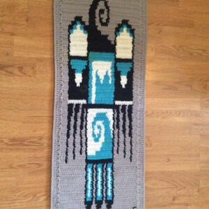 May include: A gray crocheted rug with a blue, white, and black geometric design. The rug has a fringe border and the text "5E" is visible in the bottom right corner.