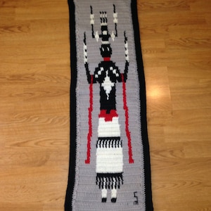 May include: A gray and black crocheted wall hanging featuring a stylized figure in black, white, and red. The figure is wearing a skirt and has long arms raised above its head. The hanging is framed with a black border.