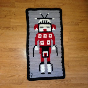 Puede incluir: Un tapiz de pared tejido a crochet con una figura nativa americana estilizada en rojo, blanco y negro sobre un fondo gris.
