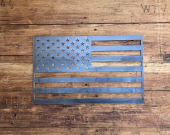 May include: A metal American flag cut-out with stars and stripes. The flag is made of a silver-colored metal and is displayed against a wooden background. The stars are cut out of the metal, and the stripes are horizontal.