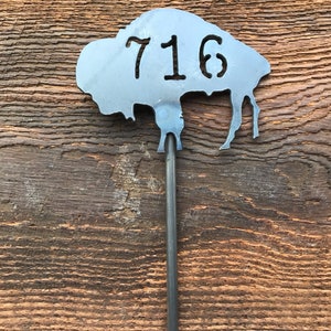 May include: Metal house number sign in the shape of a buffalo silhouette with the number 716 cut out. The sign is attached to a black metal rod.