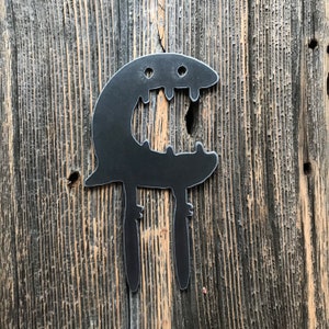 Könnte beinhalten: Ein schwarzes Metall-Wandkunstwerk in Form eines stilisierten Wesens mit offenem Mund und zwei Beinen. Das Kunstwerk ist auf einem verwitterten Holzgrund befestigt.