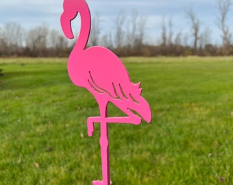 May include: Pink metal flamingo garden stake. The flamingo is standing on one leg with its head bent back. The stake is inserted into a grassy lawn.